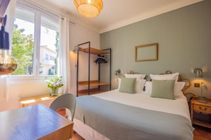 A bedroom with a bed, bedside tables, a hanging rack, a window, and a view of buildings outside. - Hôtel Les Lauriers (Saint-Tropez)