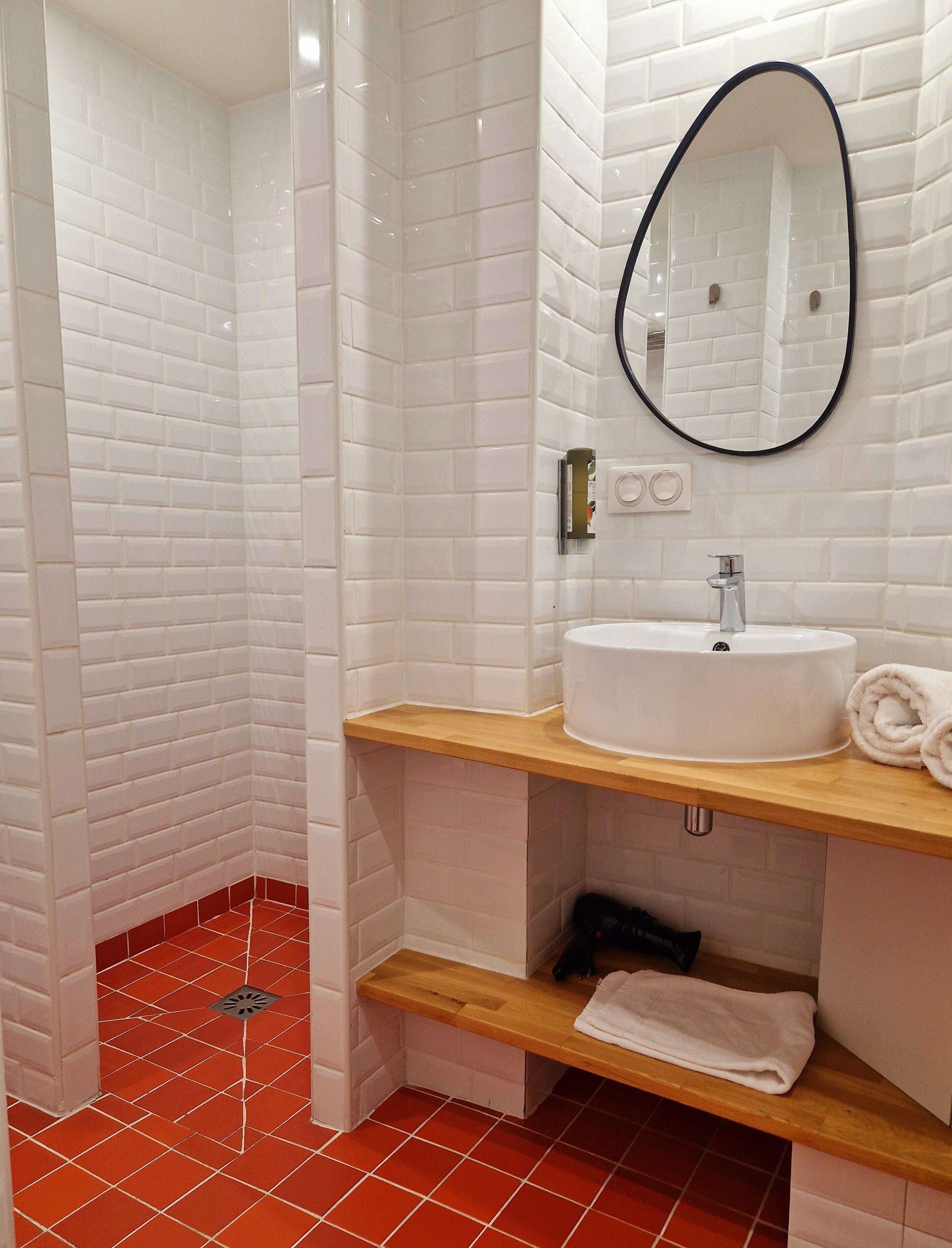 Une salle de bain avec des murs recouverts de carreaux de céramique blancs rectangulaires, un lavabo blanc avec un miroir et un sol carrelé de rouge.