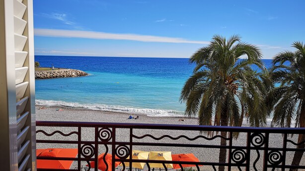 A balcony with a view of a beach, palm trees, and a rocky shoreline.