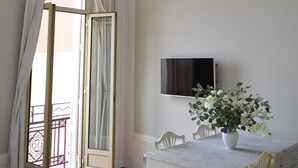 A dining area with a white table, chairs, a vase of flowers, a television on the wall, and a door leading to an outdoor balcony.