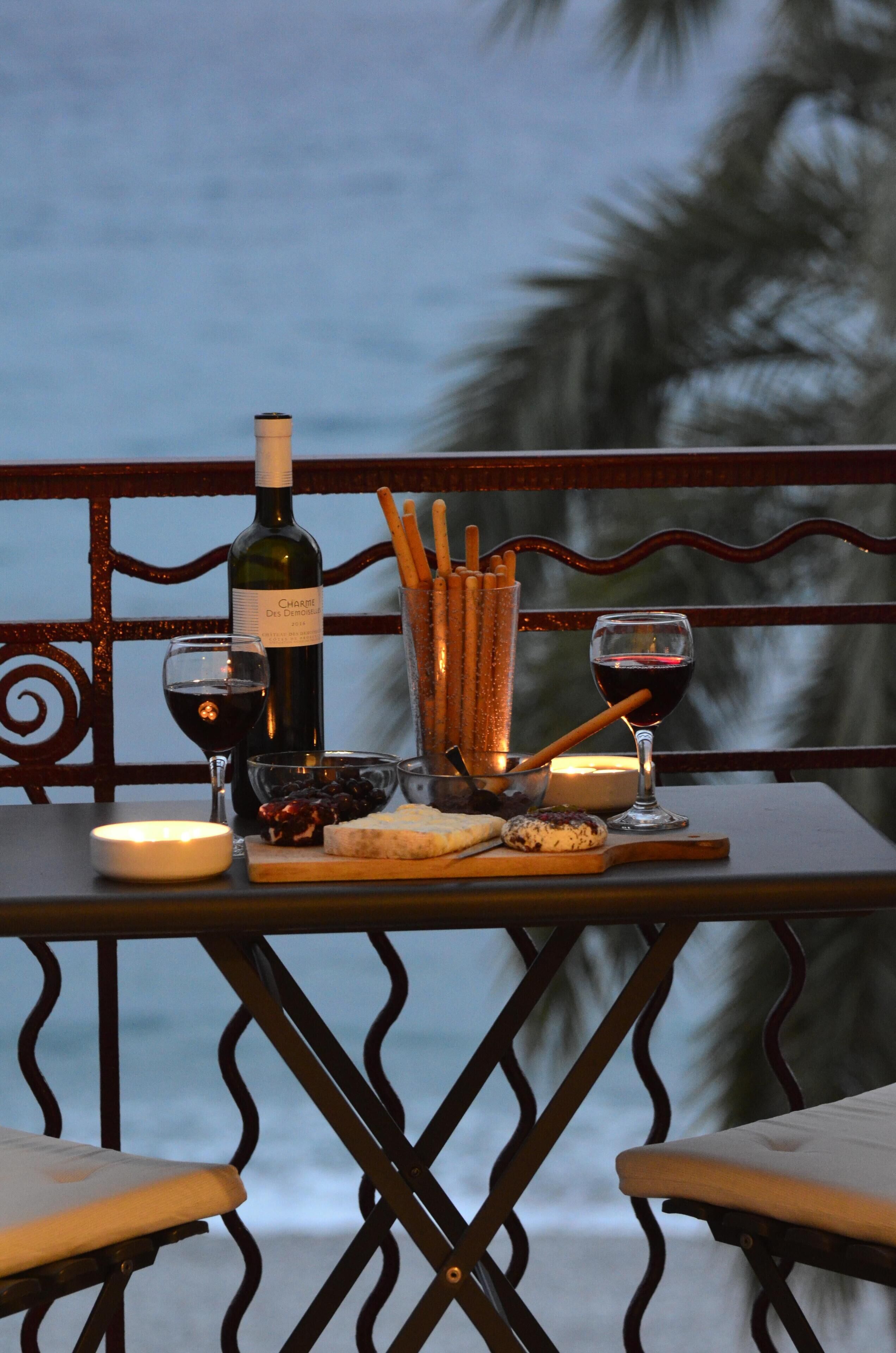 Une table dressée avec une bouteille de vin, des verres, une bougie et des amuse-gueules, sur un balcon offrant une vue sur l’océan.