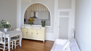 A room with a dining table, white chairs, a cream-colored cabinet, and a window with a view of a kitchen.