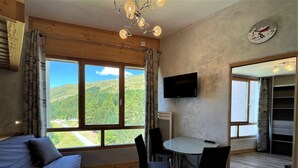 A room with a large window, a bed, a table with chairs, a TV, and a clock on the wall.