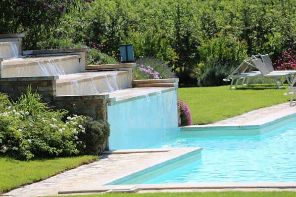 A swimming pool with a stone waterfall feature, surrounded by a well-maintained lawn and various plants. - Verdidea - Podere della Bruciata (Chiusi)