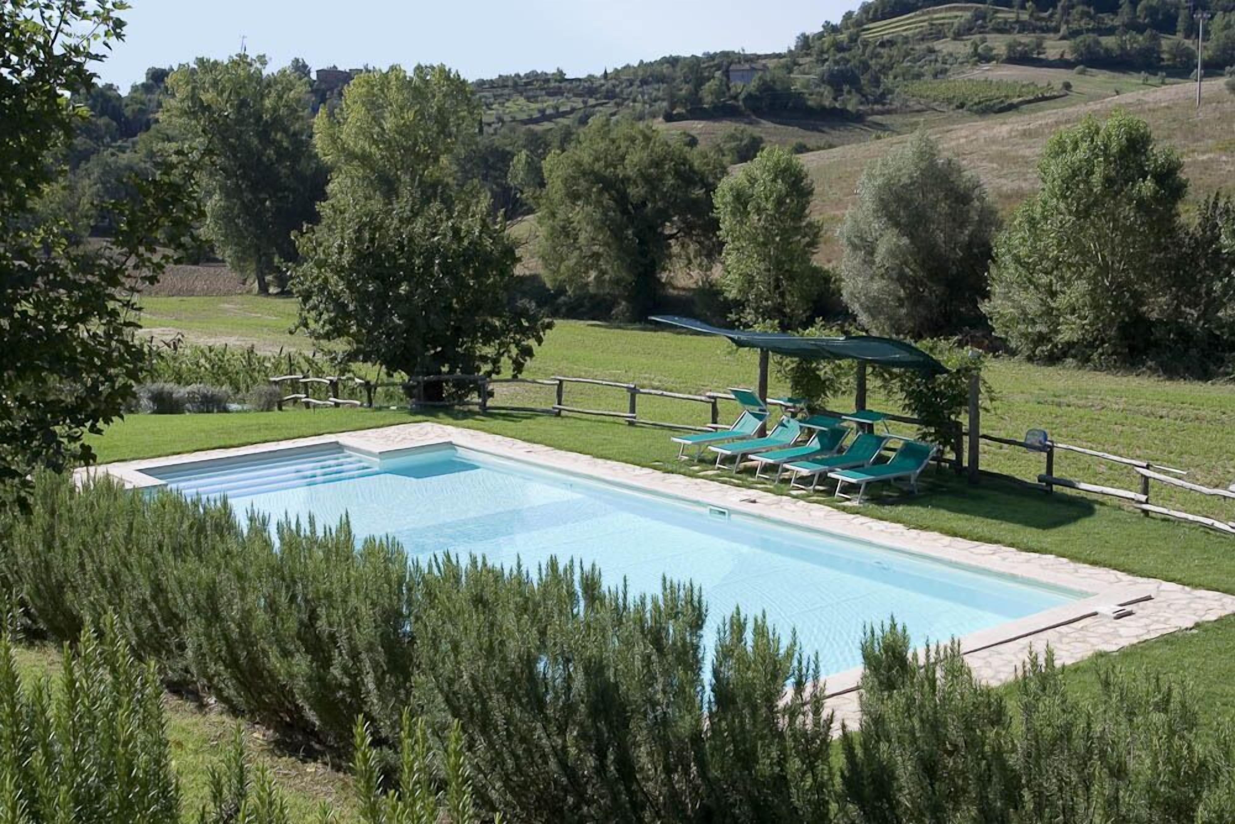 A rectangular swimming pool with a wooden deck and lounge chairs, surrounded by greenery and hills in the background.
