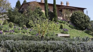 Un grand bâtiment rustique en pierre, doté d’un toit en pente, entouré d’un jardin bien entretenu où poussent diverses espèces d’arbustes et de plantes à fleurs.