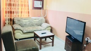 A living room with a sofa, ottoman, coffee table, TV, and a wall-mounted air conditioner. - Cossy Hotel & Suite (Agbor)