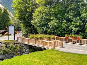 A wooden bridge over a stream with a stone wall on one side and a road on the other. There are flower planters along the bridge and a sign that reads 'Dou des Ponts'.