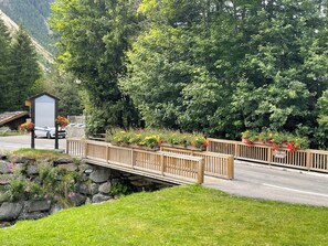 Eine Holzbrücke über einen Bach, an einer Seite eine Steinmauer und auf der anderen Seite eine Straße. Ein Schild mit Schrift in einer Fremdsprache und ein weißes Auto, das am Straßenrand geparkt ist.