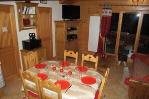 Ein Essbereich mit einem Holztisch, gedeckt für vier Personen, umgeben von Stühlen. Über dem Tisch ist ein Fernseher an der Wand angebracht.