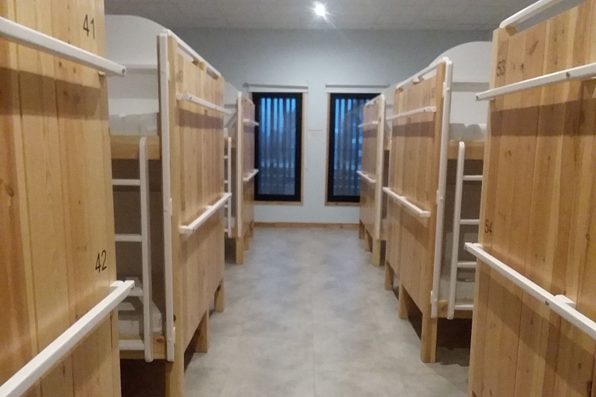 A corridor with wooden bunk beds numbered 41 to 54, leading to a room with a window and curtains.