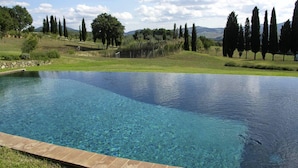 A clear blue swimming pool with a stone border, surrounded by a grassy landscape and tall trees. - Verdidea - Vallocchie (Sarteano)