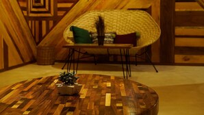 Un tavolo rotondo in legno con una piccola pianta in vaso al centro, una sedia intrecciata con cuscini e un motivo geometrico sul muro sullo sfondo.