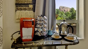 Un coin café comprenant une machine à café, un porte-tasses et des théières, le tout installé sur une table près d’une fenêtre offrant une vue sur la montagne.