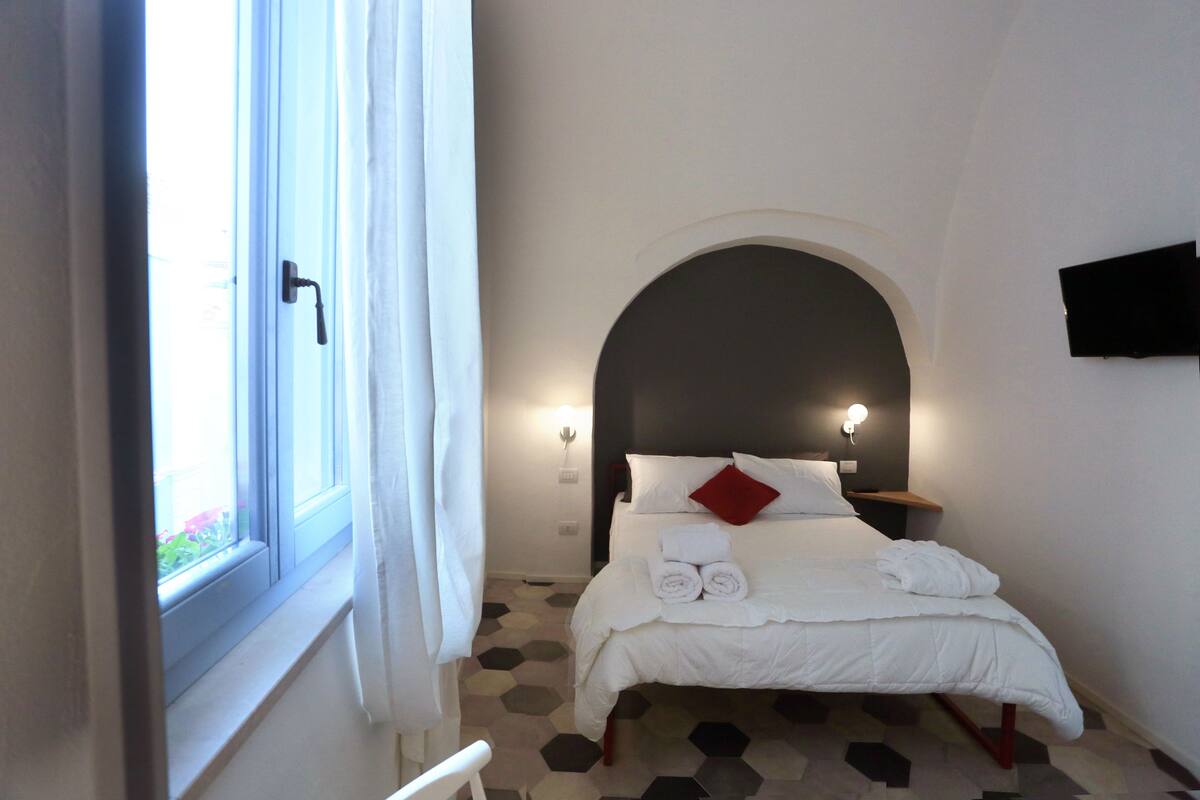 Habitación de hotel con cama, ventana con cortinas, televisor montado en la pared y suelo con estampado geométrico.