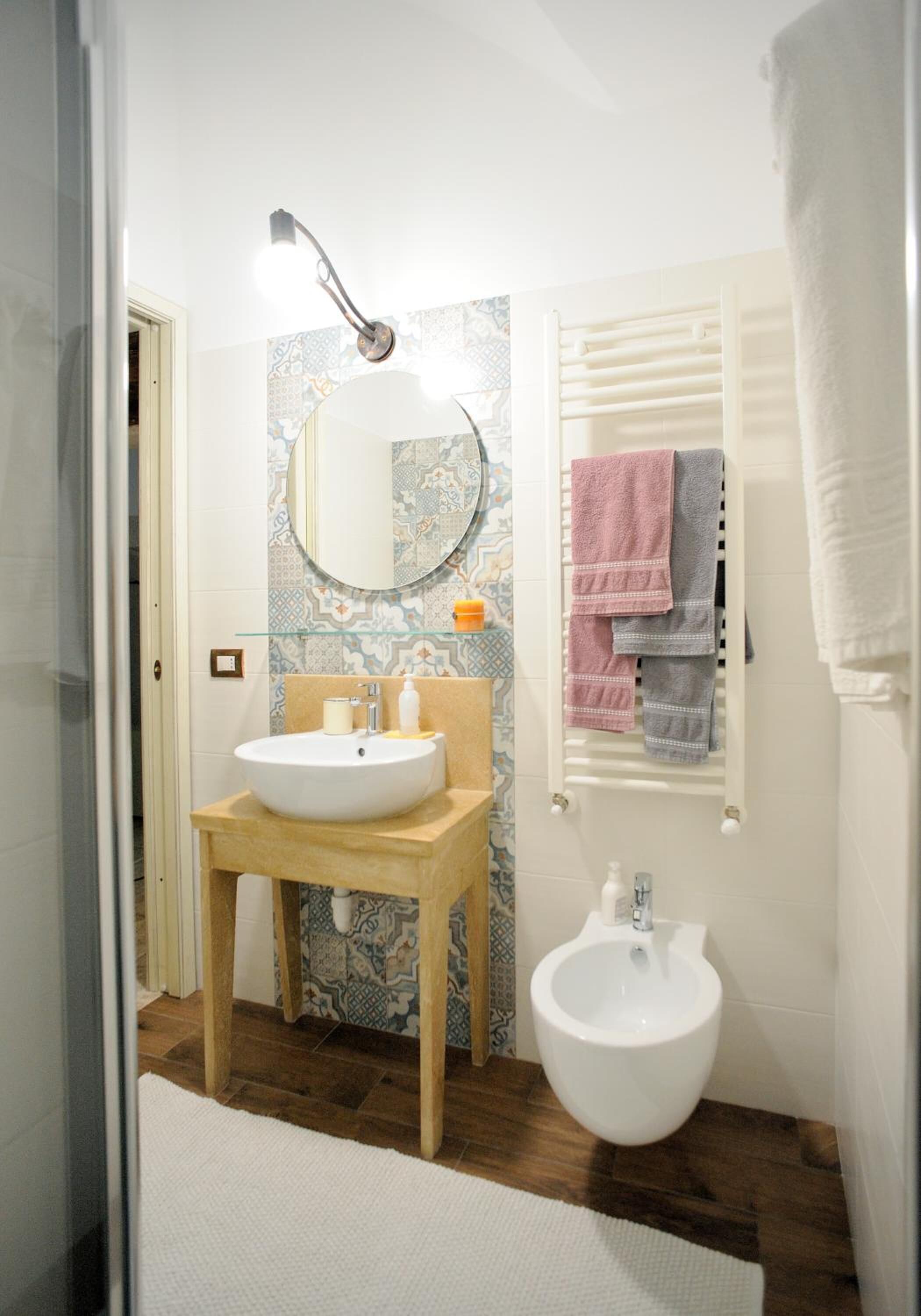 A bathroom with a wooden vanity, a white sink, a bidet, and a mirror. There are towels hanging on a rack and a glass shelf with toiletries.