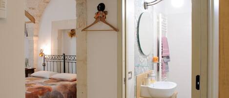 Un dormitorio con cama, viga de techo de madera, lámpara colgante y un baño con lavamanos, espejo y toallero.