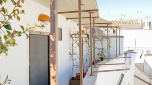 Un balcón estrecho con mesa y sillas de madera, plantas en macetas y una estructura de pérgola.