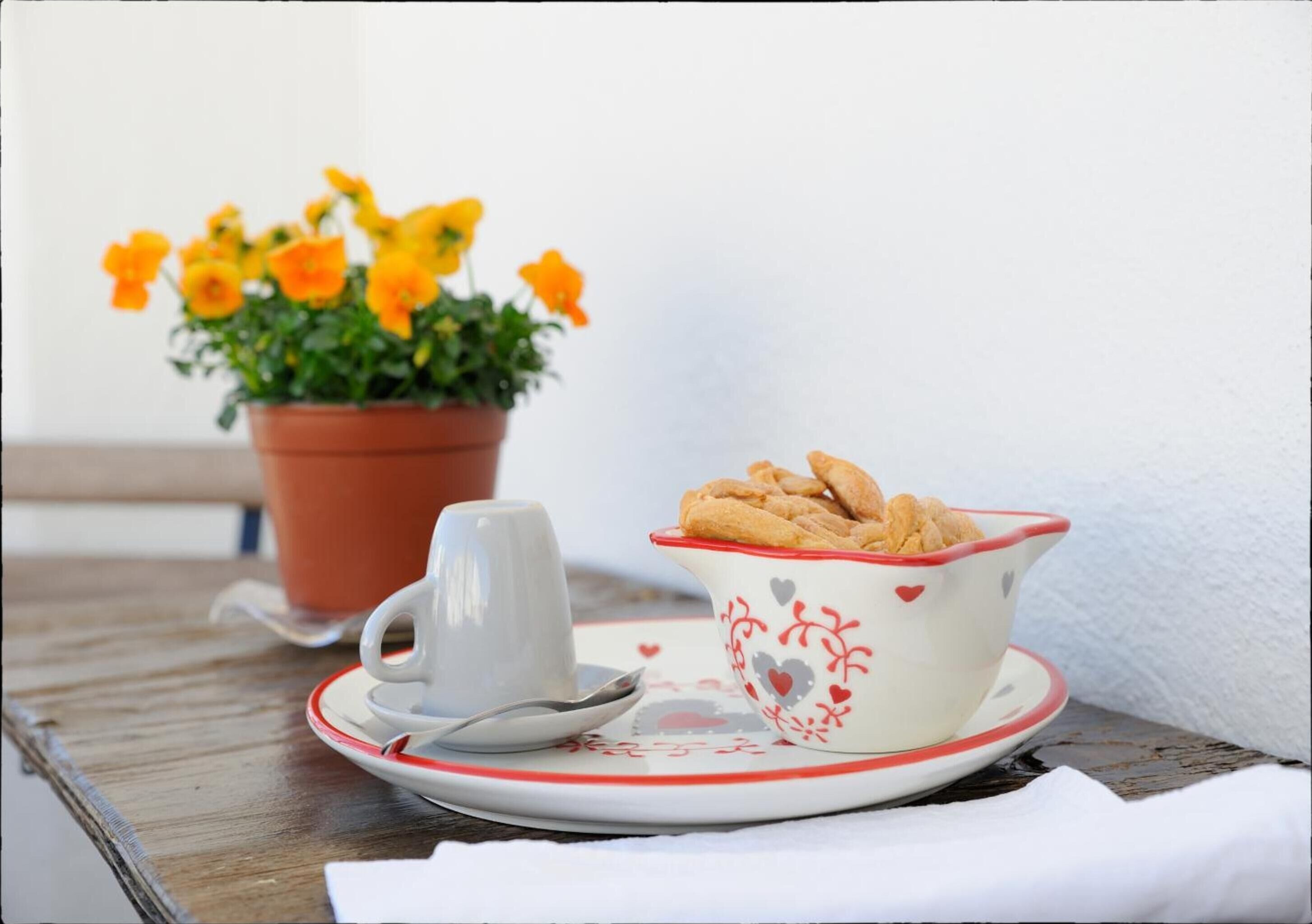 A table with a white cup and saucer, a bowl of pastries, and a potted plant with yellow flowers.