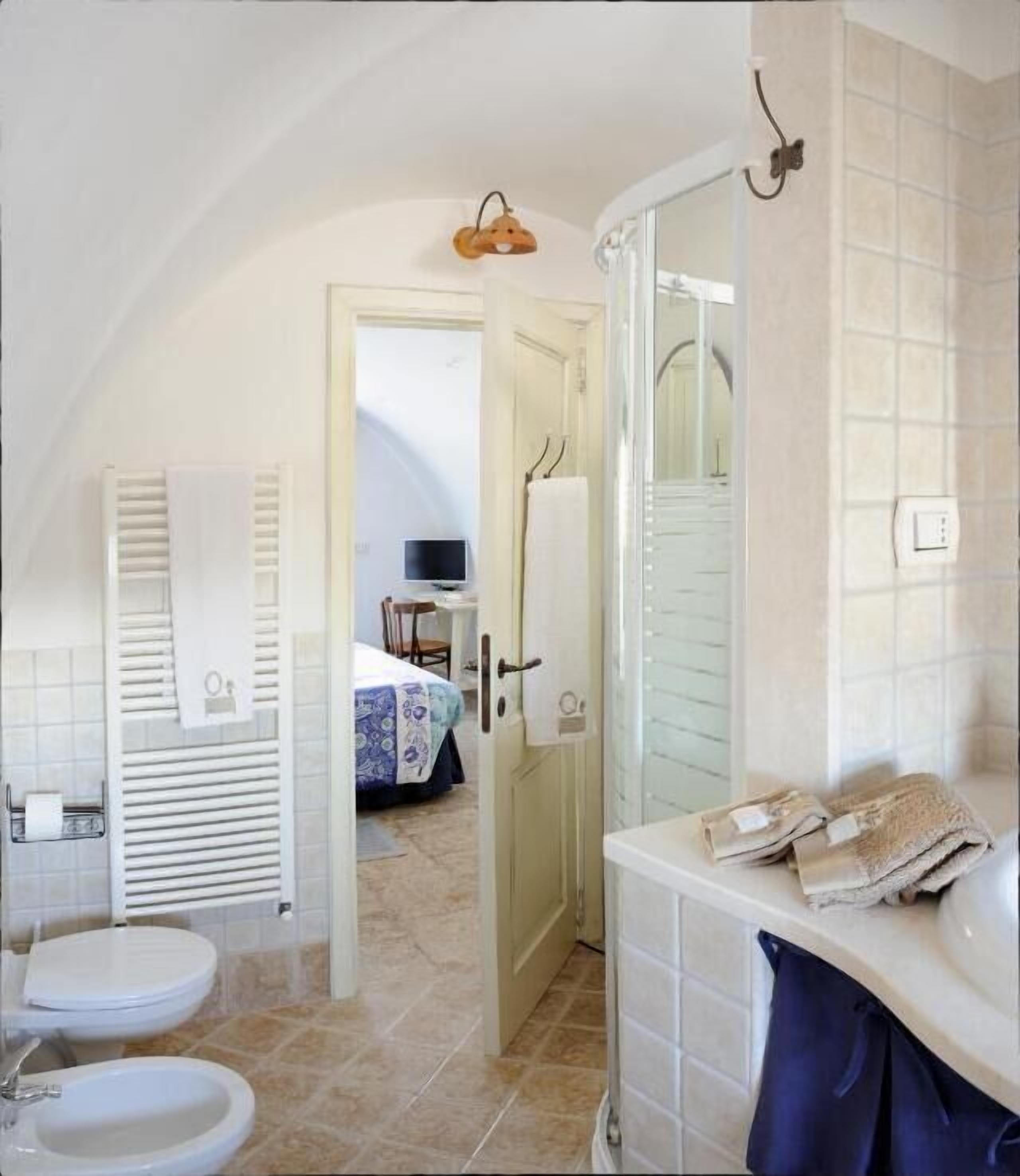 A bathroom with a bathtub, a toilet, and a sink. There is a shower area with a glass door and a towel rack. A bed is visible in the background.