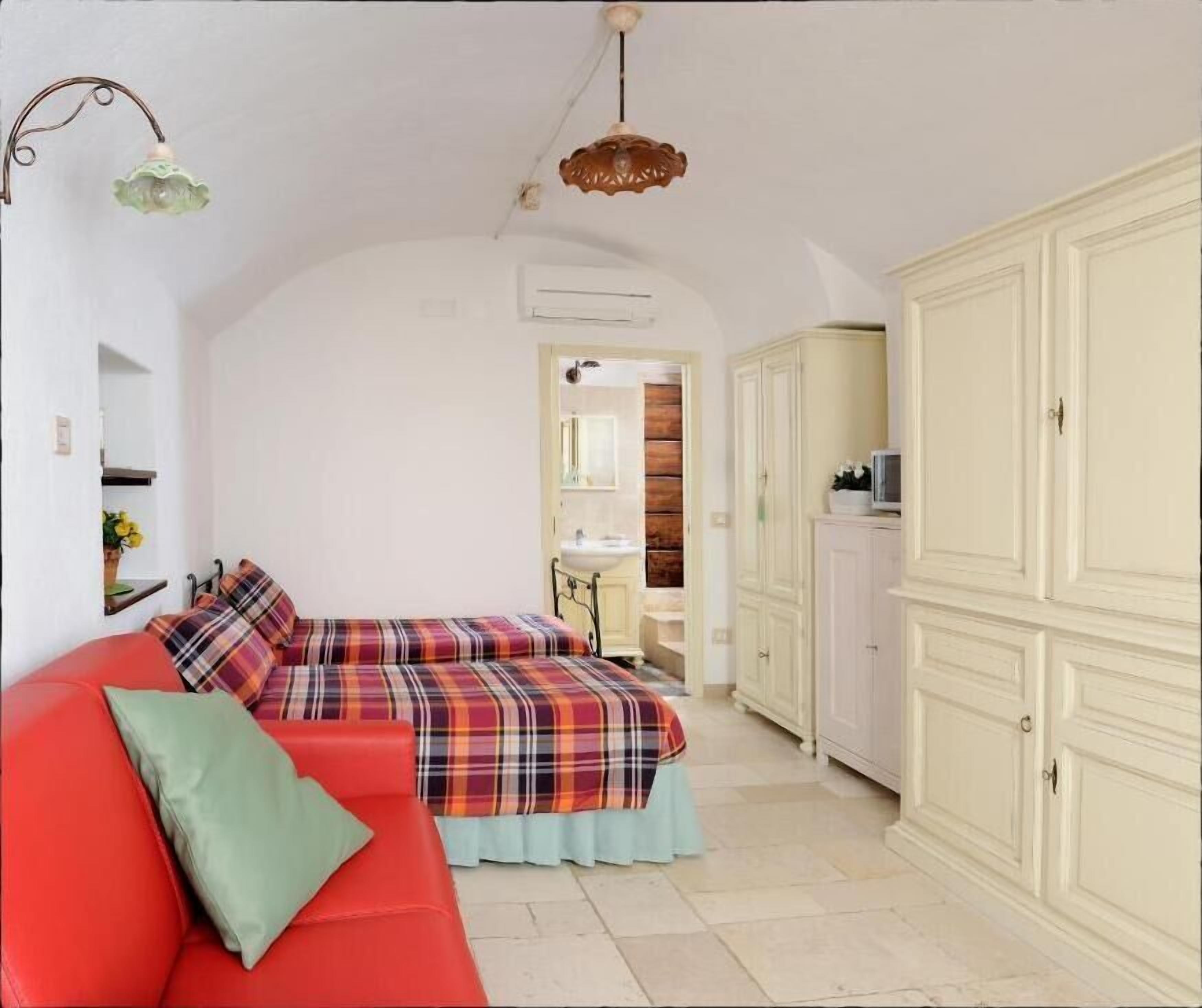 A room with a red sofa, a plaid bedspread, a white wardrobe, and a small bathroom with a sink and mirror.