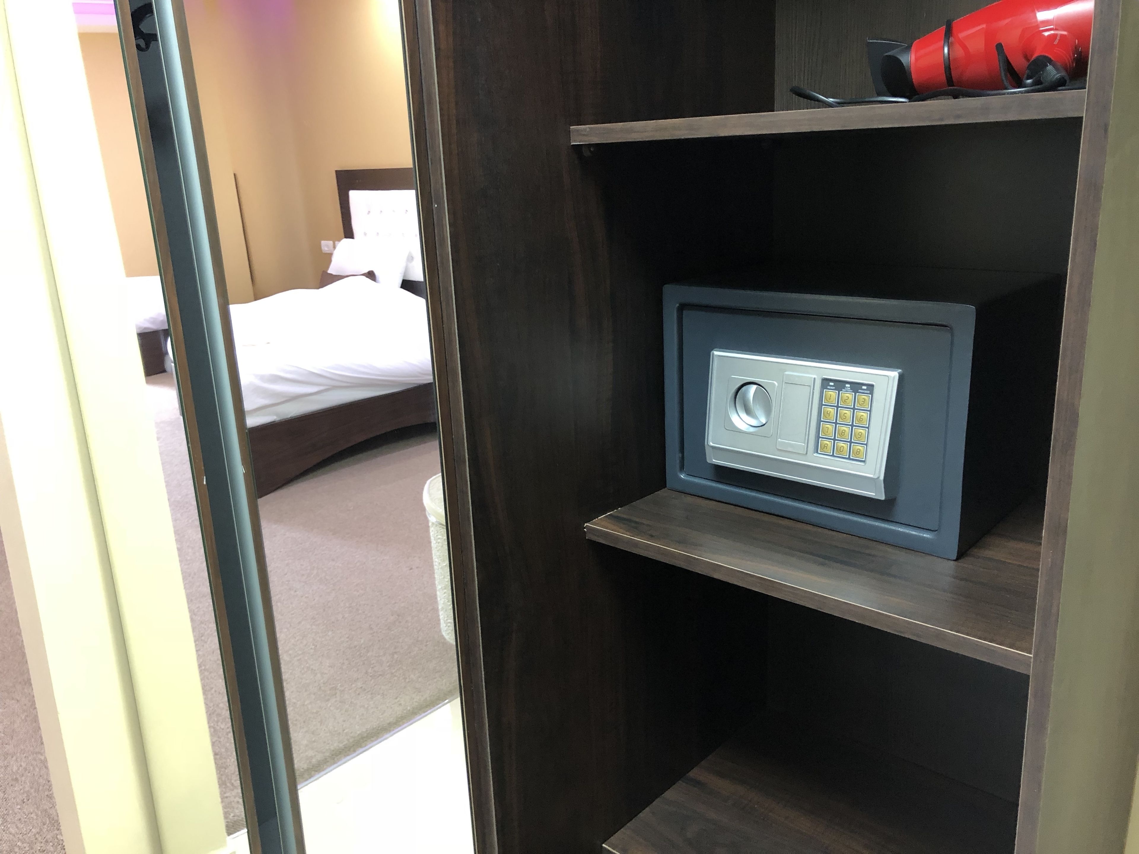 A hotel room with a bed, a safe, and a red suitcase on a shelf.
