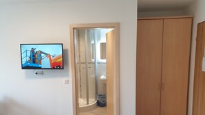A hotel room with a bed, a television mounted on the wall, a wooden wardrobe, and a glass door leading to a bathroom. - Gasthaus und Pension Stanek (Hofkirchen)