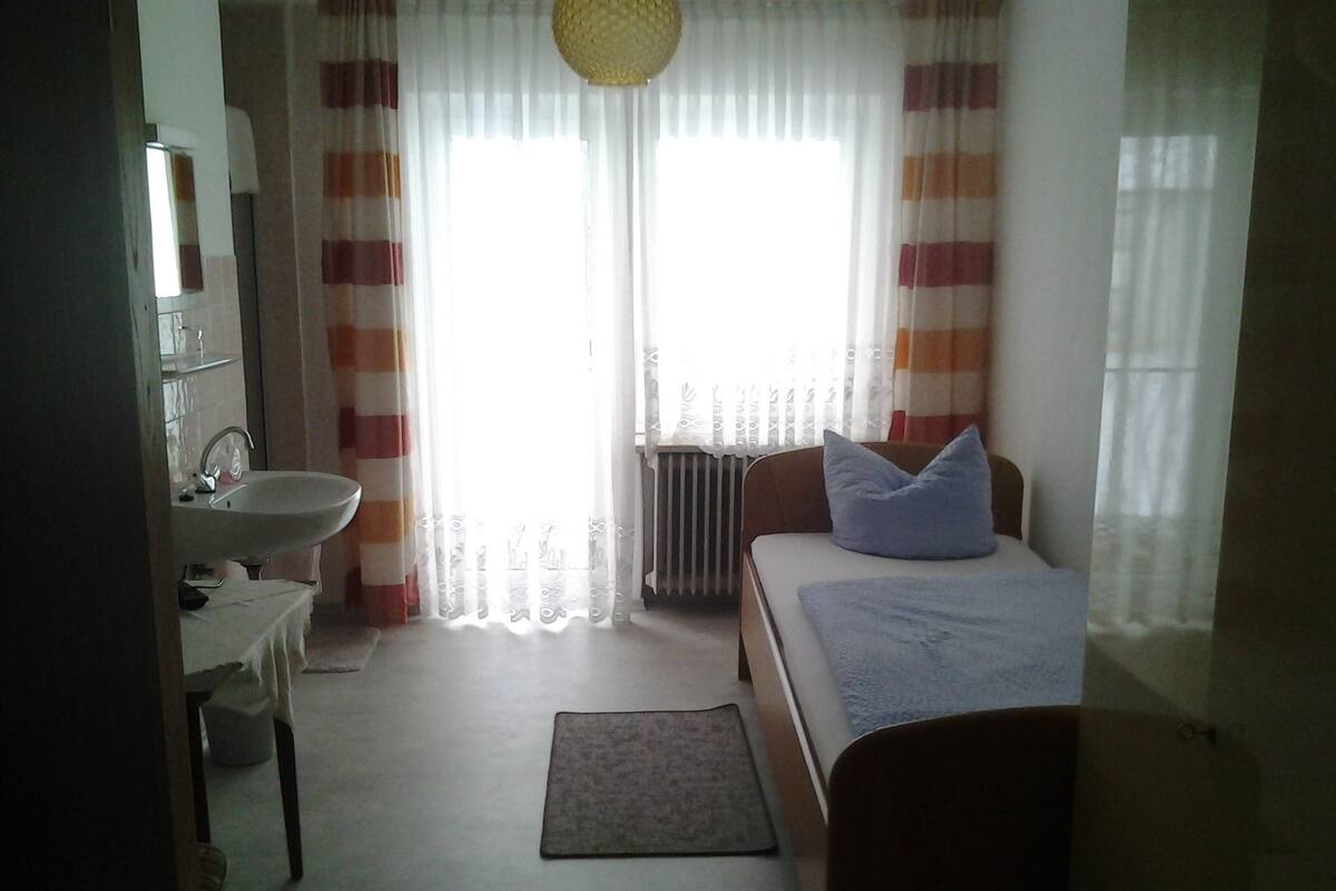 A bedroom with a bed, a bedside table, a lamp, a window with curtains, and a radiator.