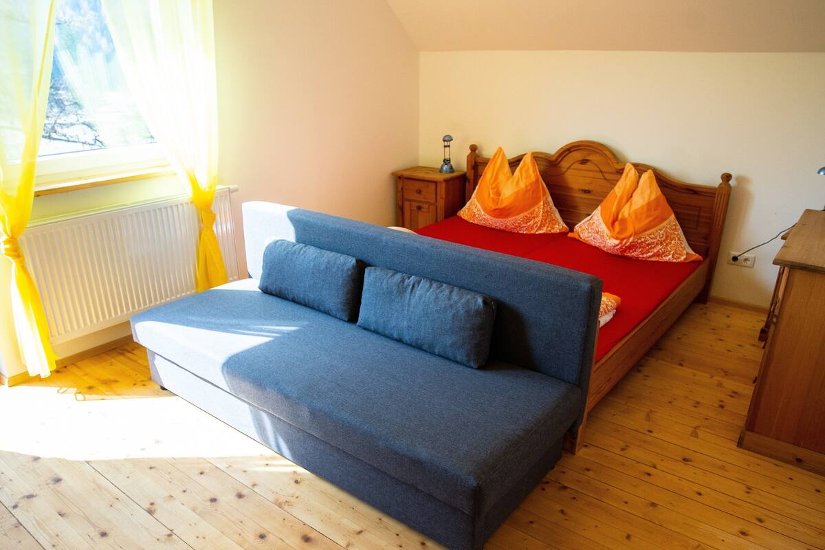 Um quarto com um sofá azul, uma cama de madeira com roupa de cama vermelha e uma mesa de cabeceira de madeira com uma lâmpada.