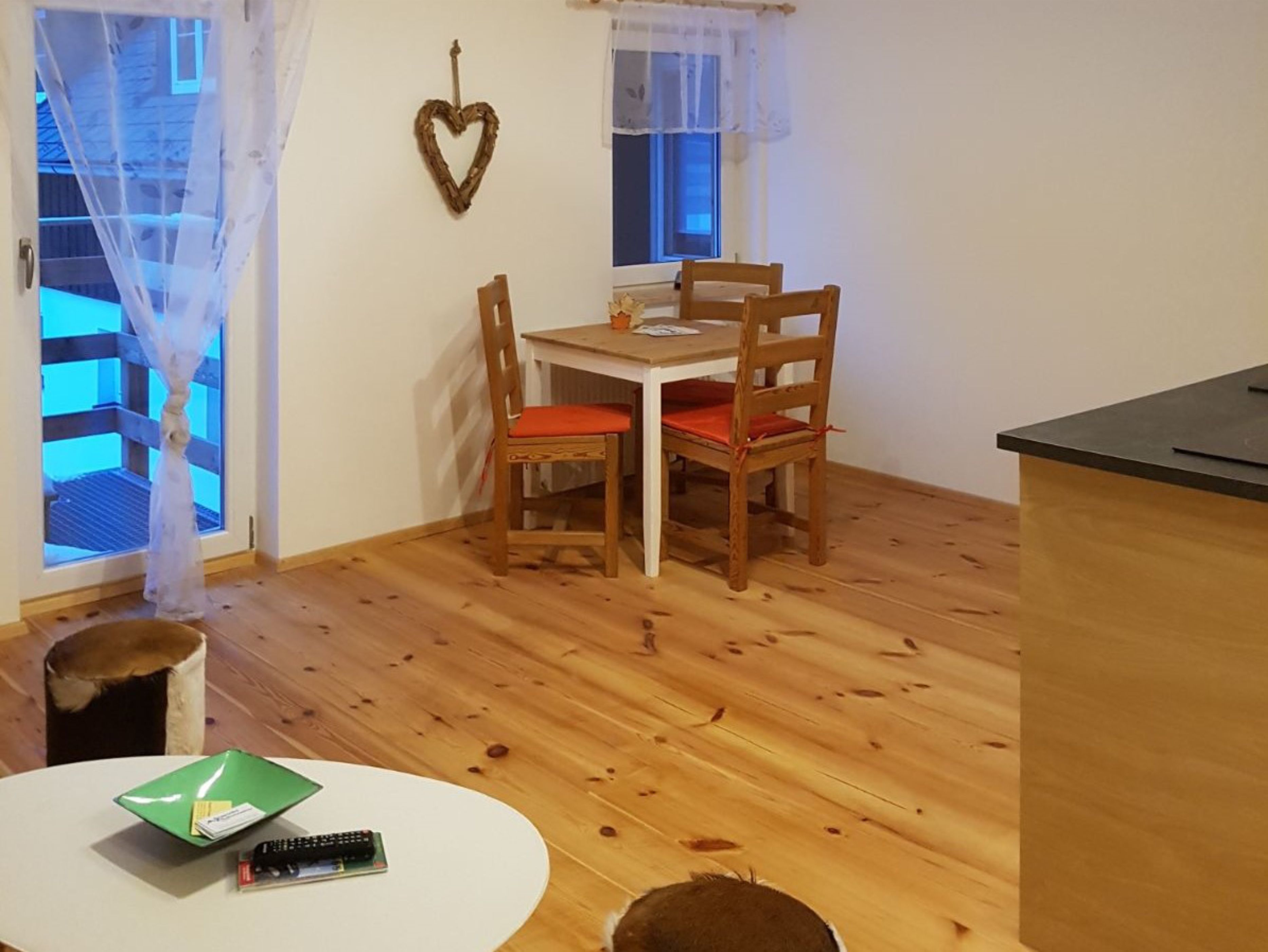 A small dining area with a wooden table and chairs, a heart-shaped decoration on the wall, and a window with sheer curtains.