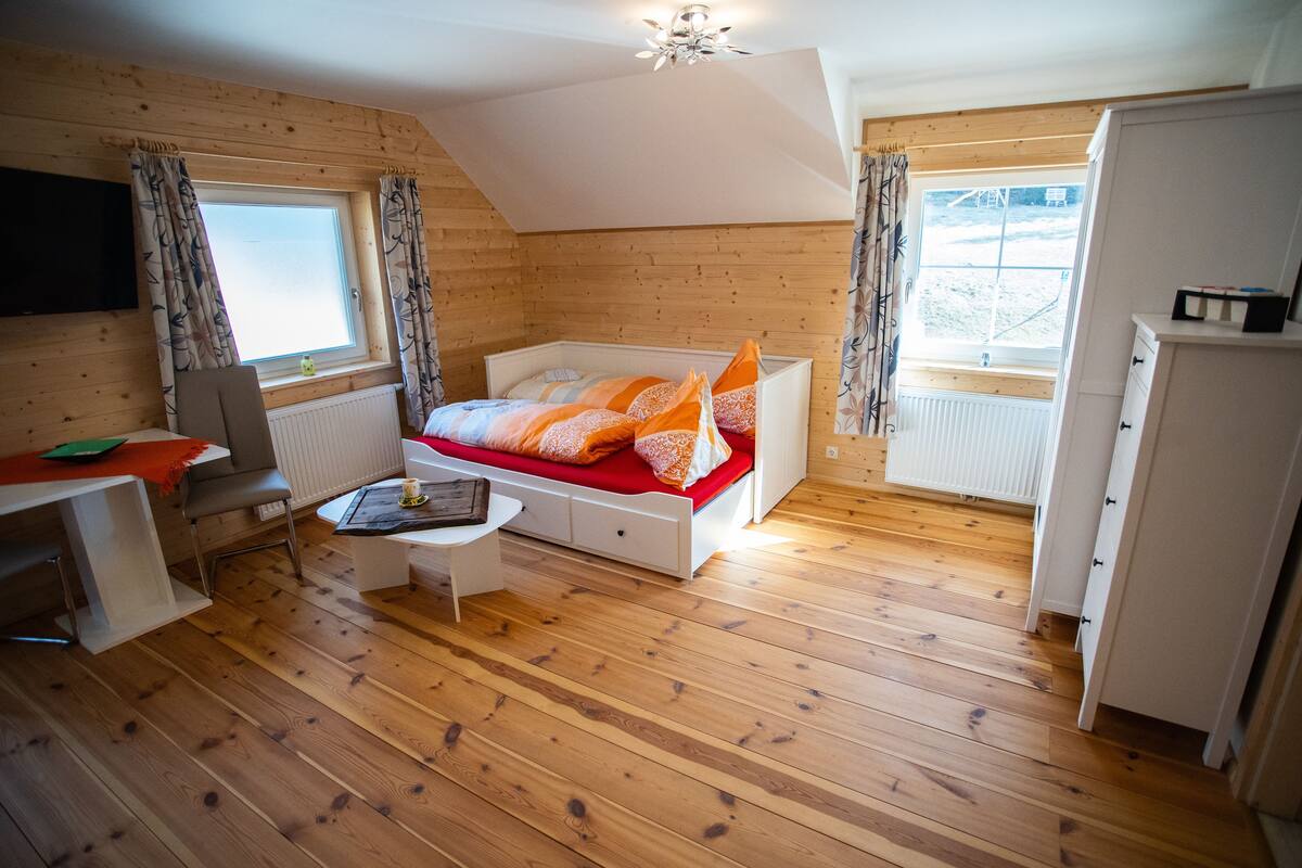Quarto aconchegante com uma cama, uma escrivaninha e uma cadeira. Há uma janela com cortinas, um radiador e um ventilador de teto.