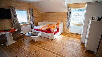 Ein gemütliches Zimmer mit einem Bett, einem Schreibtisch und einem Sessel. Es gibt ein Fenster mit Vorhängen, eine Heizung und einen Deckenventilator.