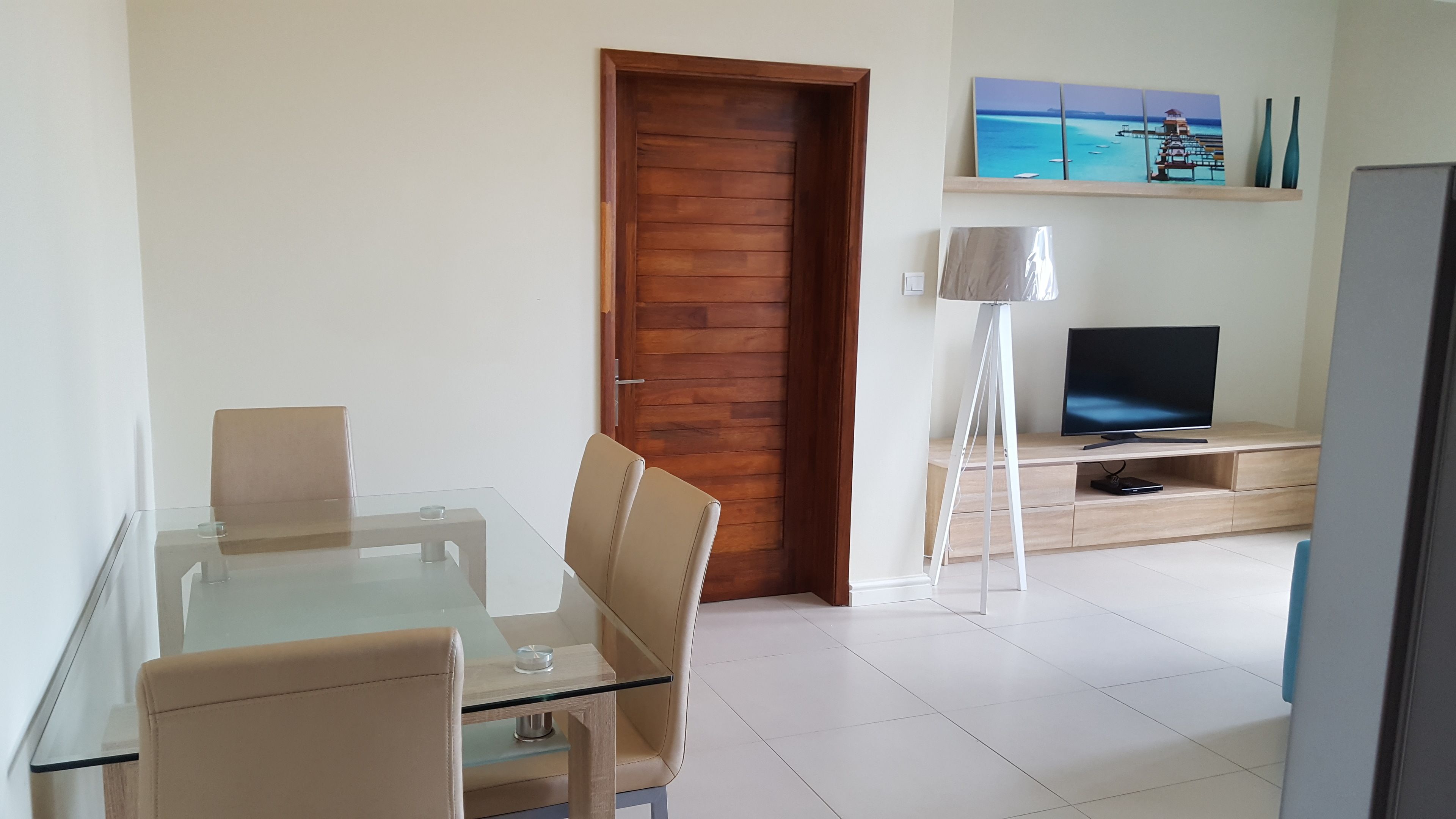 A modern living room with a glass dining table, beige chairs, a wooden door, a flat-screen TV, and a floor lamp.