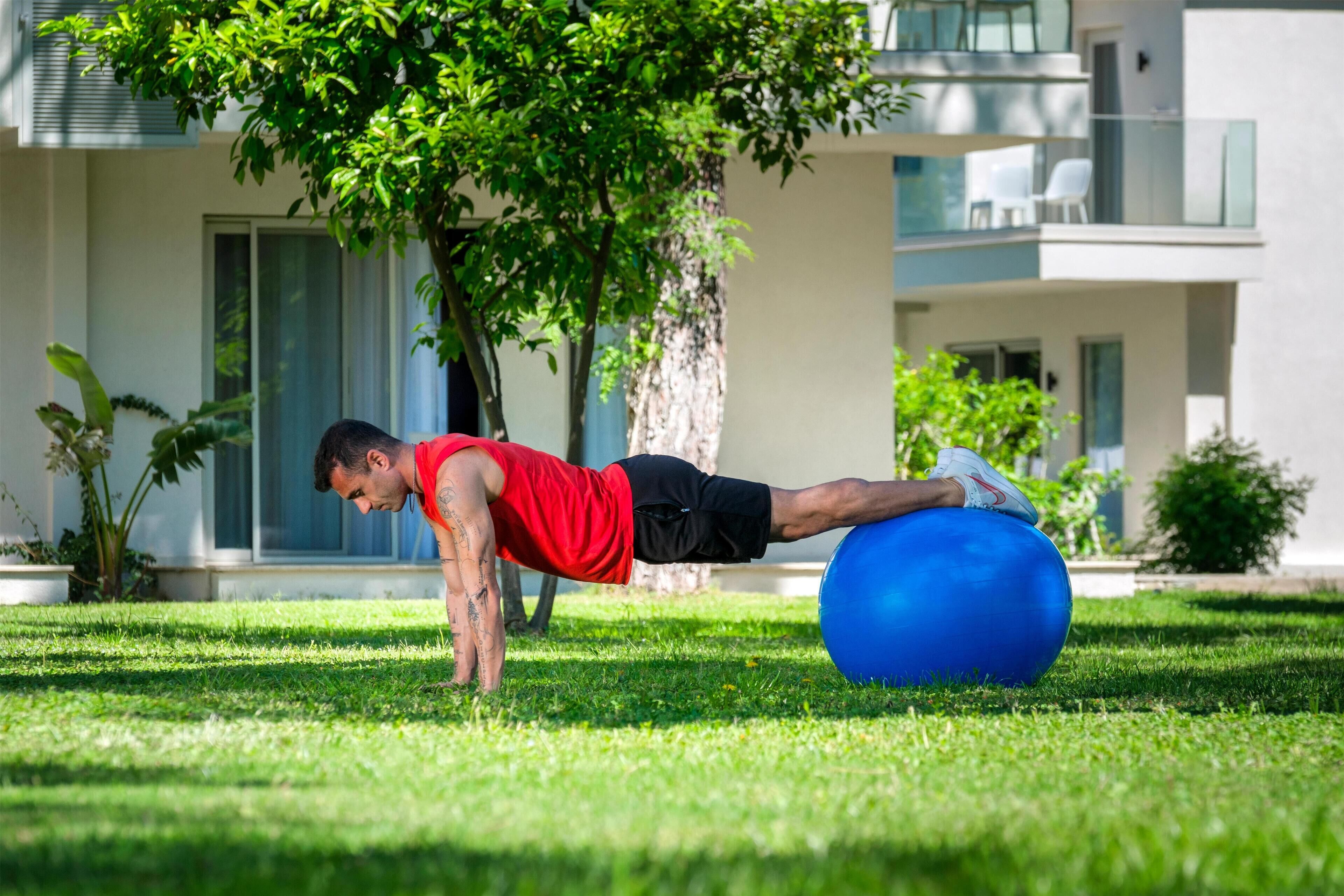 A man doing a plank exercise on a blue exercise ball in a grassy area with a building in the background.