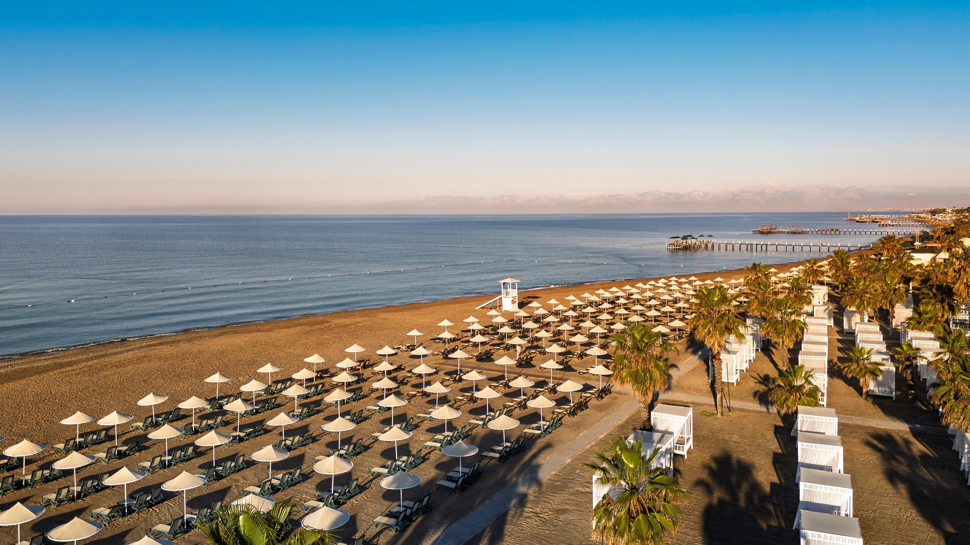 A beach resort with rows of sun loungers and umbrellas, a lifeguard tower, and a pier extending into the sea.