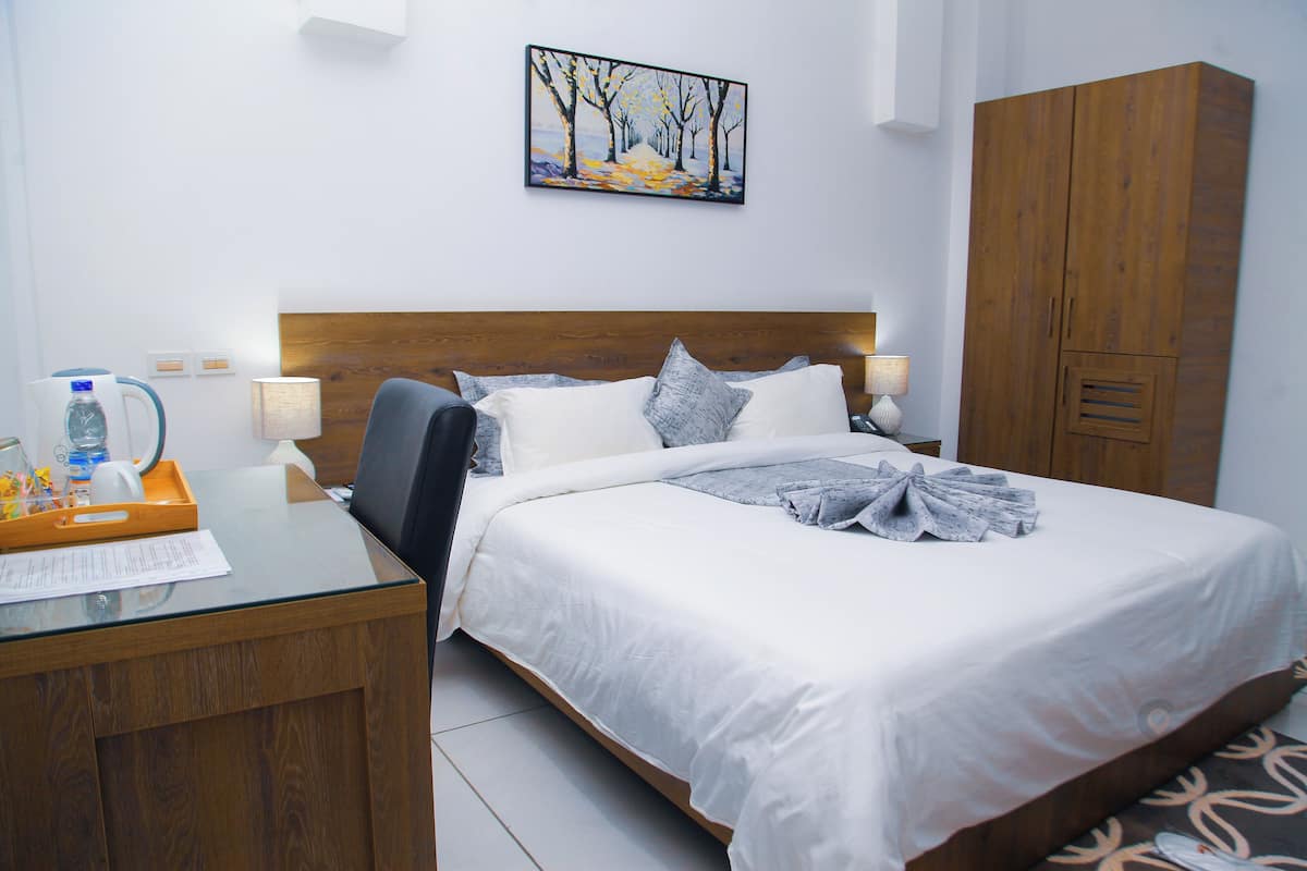 A hotel room with a wooden headboard, a bed with white bedding, a desk with a tray of items, a lamp, and a painting on the wall.