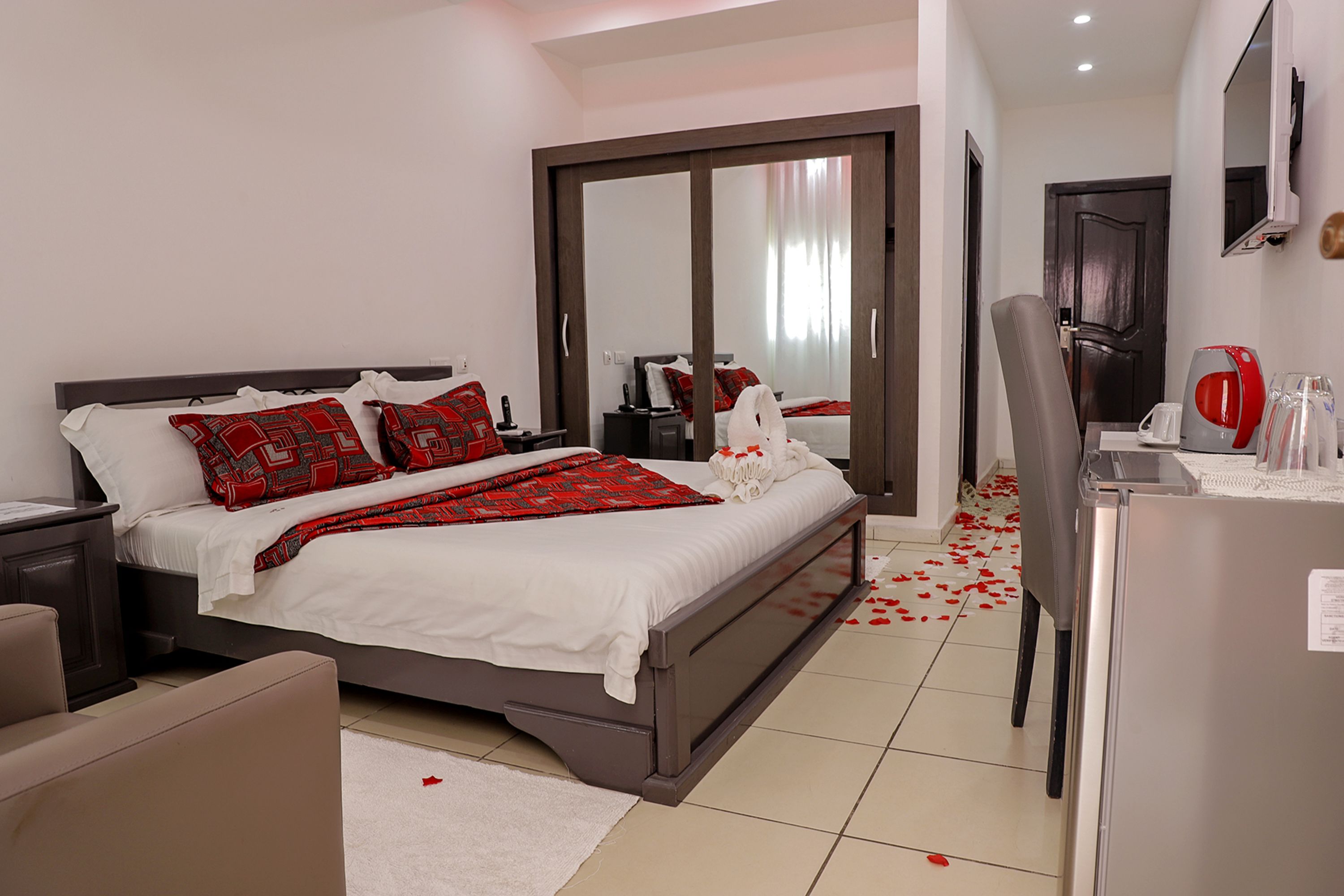 A modern hotel room with a large bed, a desk with a refrigerator, and a mirror.