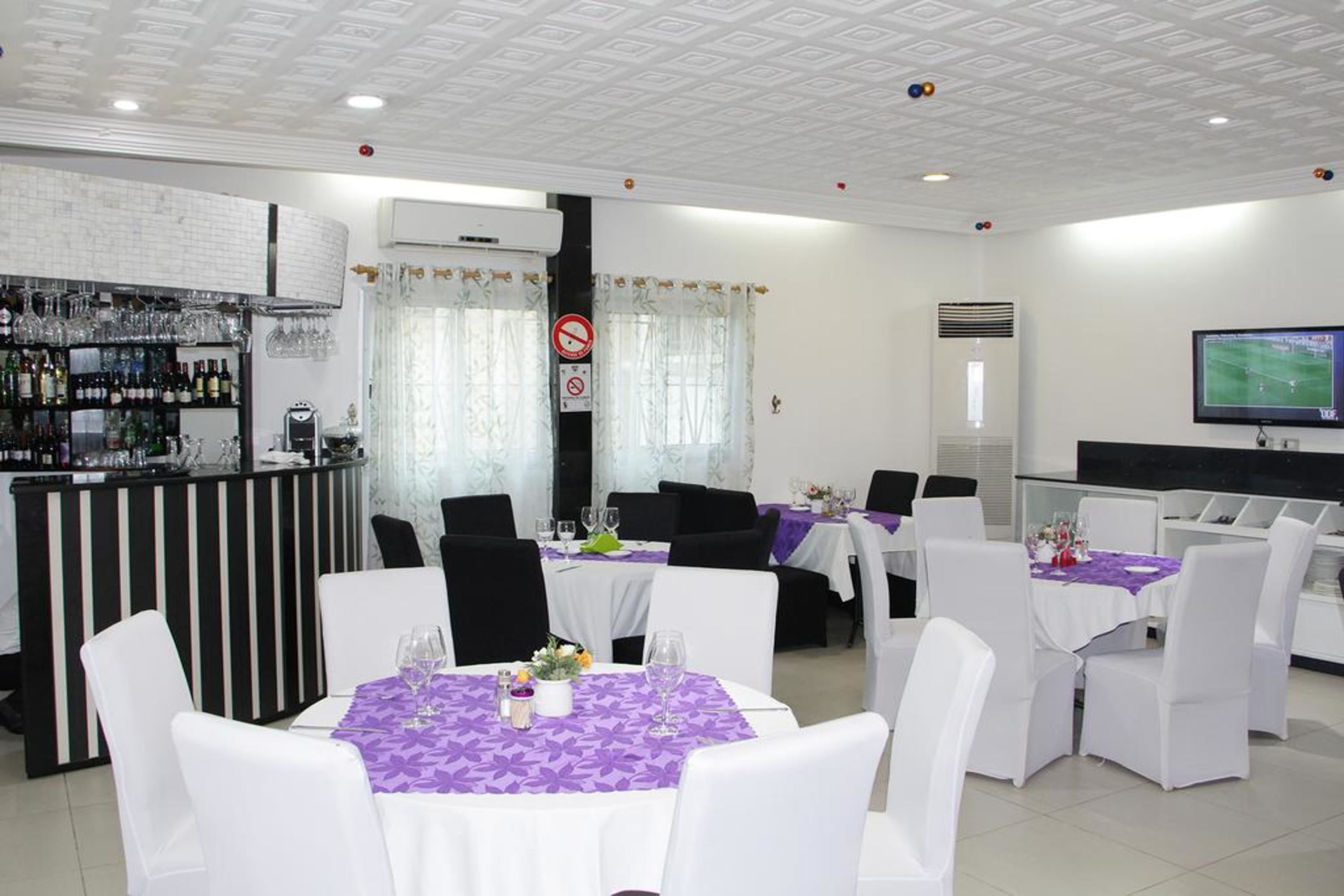 A restaurant with white chairs and purple tablecloths, a bar area with bottles, and a TV screen on the wall.