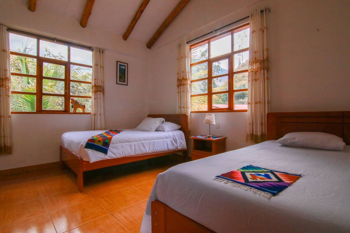 A room with two beds, wooden floors, and large windows with curtains.