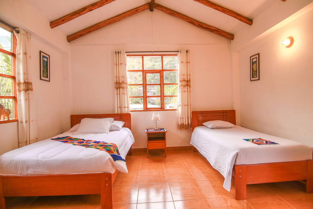 A room with two wooden beds, a small nightstand, a window with curtains, and a wall-mounted light.