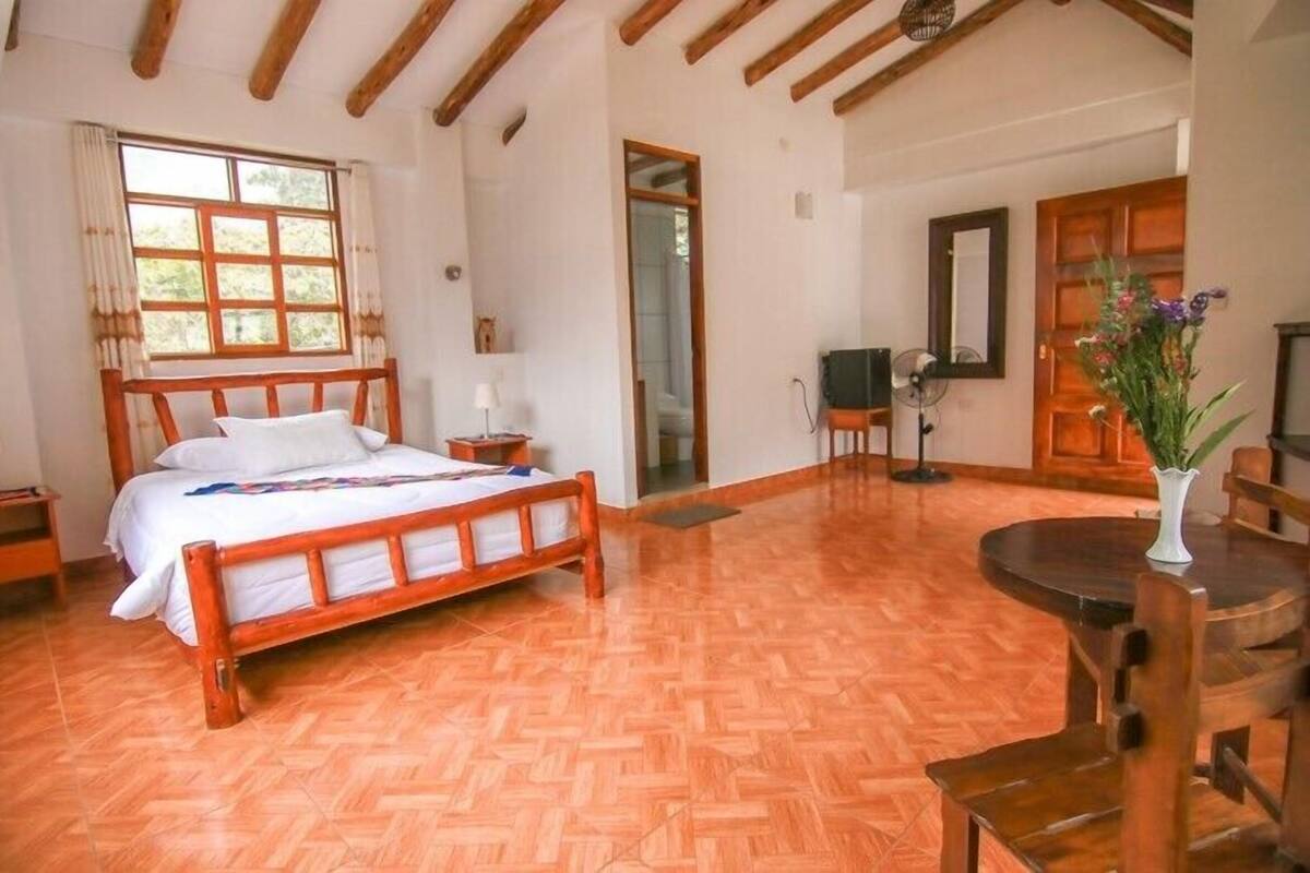 A bedroom with a wooden bed, a wooden table, and a chair. There is a window with curtains and a door in the background.
