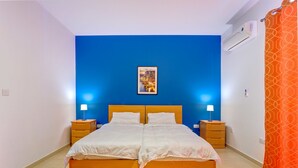 A double bed with white bedding, two bedside tables with lamps, a framed picture on the wall, and an air conditioning unit.