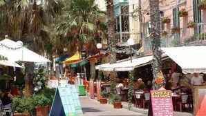 A street with outdoor seating at cafes, palm trees, and a sign for 'CHEZ GAETANE'. - Luxury Holiday Apartment IN Qawra (St. Paul's Bay)