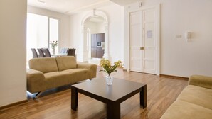 Una sala de estar moderna con un sofá color beige, una mesa de centro y un florero con flores. En el fondo, se ve un comedor con una mesa y sillas.