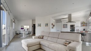 A modern living room with a beige sectional sofa, a dining area with a white table and chairs, and a kitchen with stainless steel appliances. - Luxury 2 Bedroom Penthouse in St Julians (Swieqi)