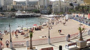 A beach with people sunbathing and swimming, a promenade with benches, and buildings in the background. - Luxury 2 Bedroom Penthouse in St Julians (Swieqi)