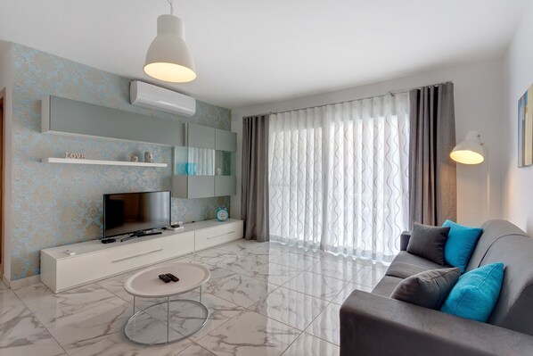 A modern living room with a flat-screen TV, a white entertainment unit, a marble-tiled floor, a grey sofa with blue cushions, and a large window with sheer curtains. - Luxury 2 Bedroom Apartment by the Sea (St. Paul's Bay)