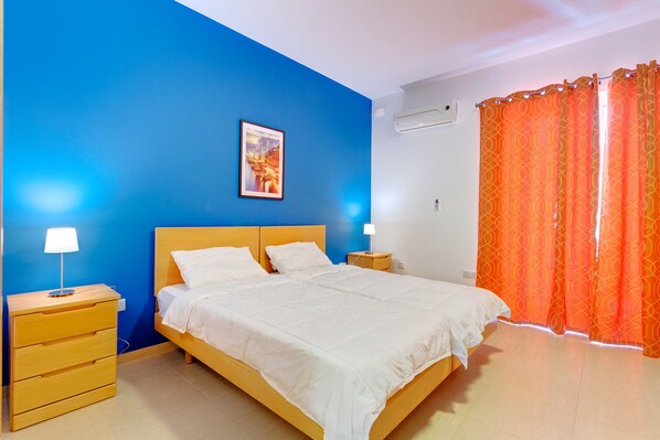 A bedroom with a bed, two bedside tables, a nightstand, a wall-mounted air conditioner, and a window with orange curtains.
