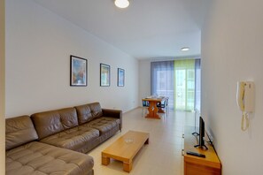 A modern living room with a brown leather sofa, a wooden coffee table, and a dining area with a table and chairs. There are framed pictures on the wall and a telephone on the wall.