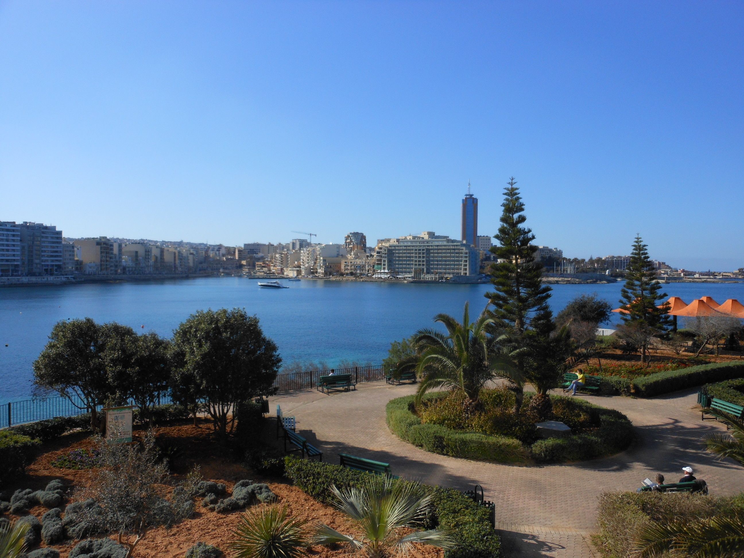 A coastal cityscape with a marina, a prominent tall building, and a park with benches and pathways.